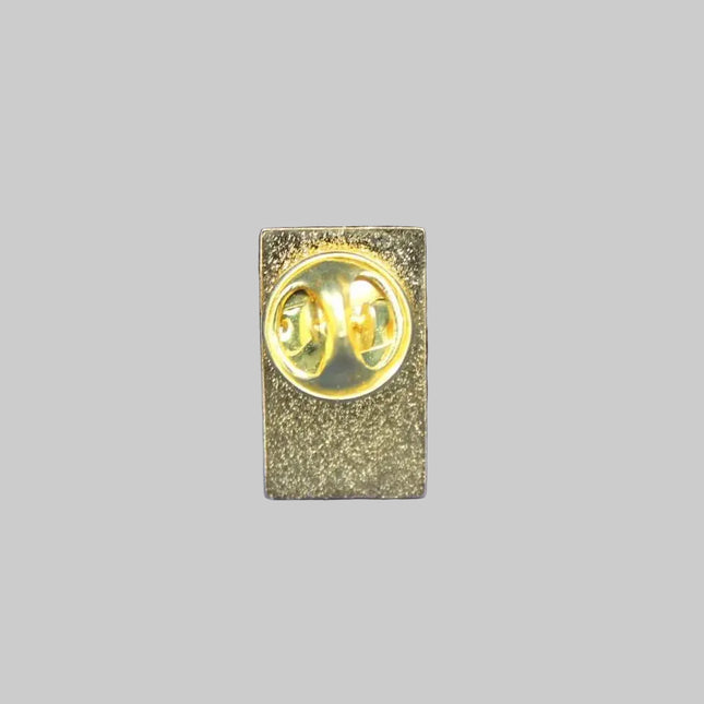 A small gold-toned rectangular pin or badge with a circular emblem in the center, placed on a textured blue fabric background.