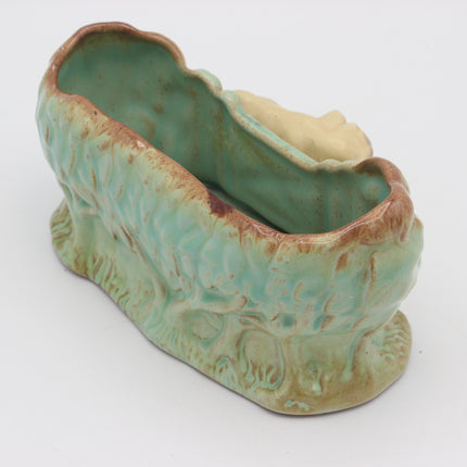 A ceramic nursery cradle or doll bed with a textured, organic shape, featuring a glossy light green and turquoise glaze with brown distressed accents and a cream-colored opening.