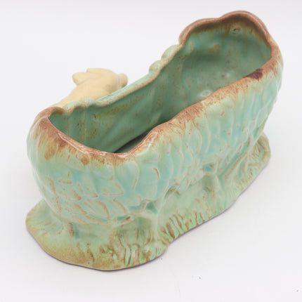 A vintage ceramic nursery planter shaped like a rustic wooden manger, glazed in a light green and beige color with a small beige figurine inside.