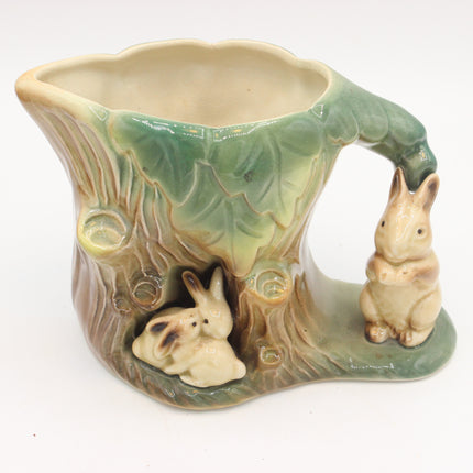 A ceramic mug shaped like a tree trunk with a handle, featuring two rabbit figures and a green leafy rim, photographed on a plain white background.