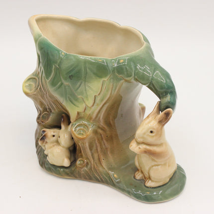 A ceramic planter shaped like a tree stump with rabbit figures, featuring a glossy glaze in shades of green, beige, and brown on a white background.