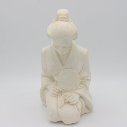 An off-white a alabaster figurine of a kneeling figure in traditional robes holding a round fan, set against a seamless light gray background. 