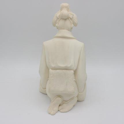 An off-white alabaster  figurine of a kneeling figure seen from the back, set against a seamless light gray studio background.