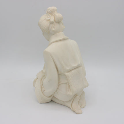 An off-white alabaster  figurine of a kneeling woman seen from behind, wearing a kimono-style robe with her hair in a bun, set against a seamless light gray background.