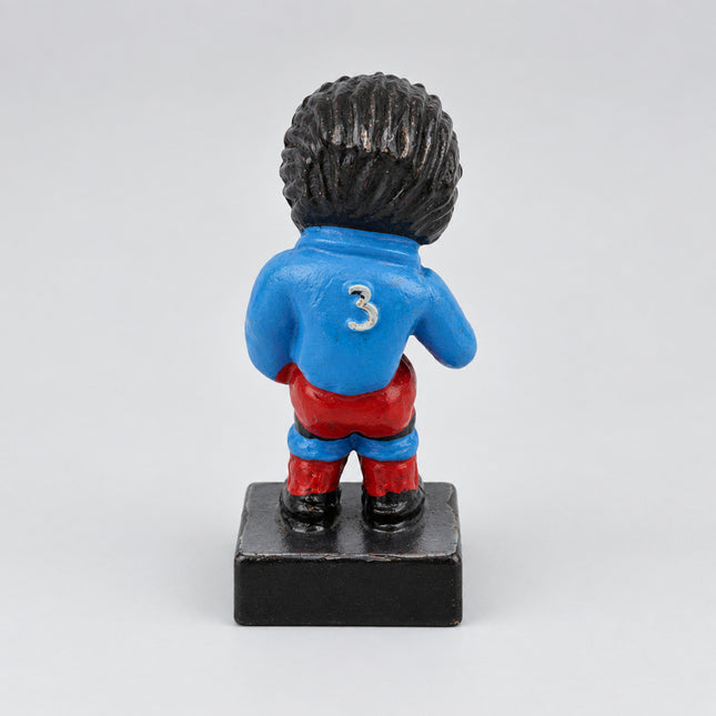 A small painted Golly figurine shown from the back on a plain light background. The figure has black hair, a blue top with a white number 3, red pants, and a dark base.
