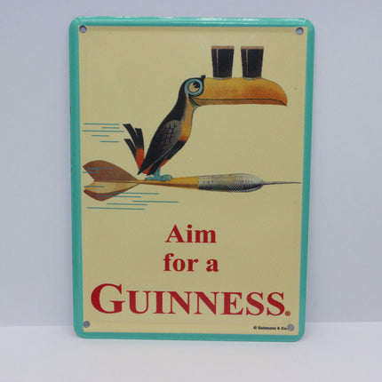 Vintage Guinness tin sign featuring a toucan balancing two pint glasses on its beak while perched on a dart, with the text 'Aim for a GUINNESS' on a cream background with a turquoise border.