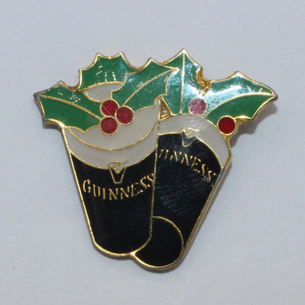 A small enamel pin shaped like two Guinness cans topped with green holly leaves and red berries, shown on a plain light background.