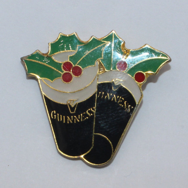 A small enamel pin shaped like two Guinness cans topped with green holly leaves and red berries, shown on a plain light background.