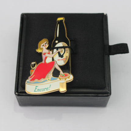 Enamel pin with a woman in a red dress and a bottle labeled 'Encore!' in a black box.