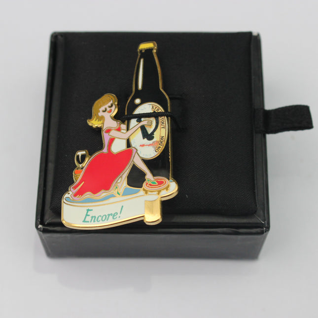 Enamel pin with a woman in a red dress and a bottle labeled 'Encore!' in a black box.