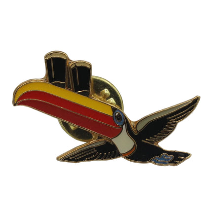 A small enamel pin shaped like a toucan with a gold-tone metal outline, featuring black, red, yellow, and white coloring, photographed on a plain light gray background.