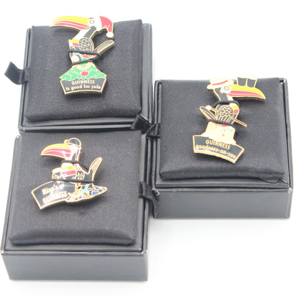 Three enamel lapel pins displayed on black presentation boxes against a white background. The pins feature toucan and guinea bird designs with 'GUINNESS' and 'Black Guineas' text.