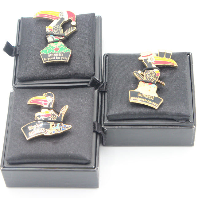 Three enamel lapel pins displayed on black presentation boxes against a white background. The pins feature toucan and guinea bird designs with 'GUINNESS' and 'Black Guineas' text.