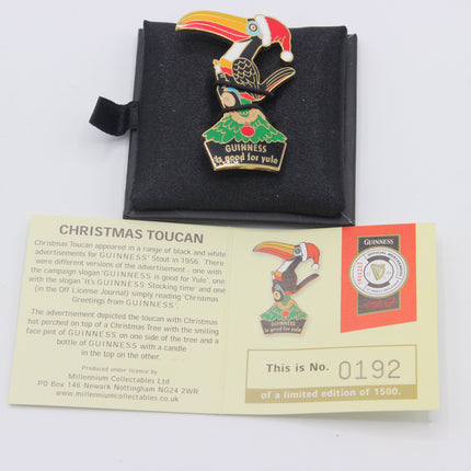 A Guinness Christmas Toucan enamel pin displayed on a black velvet stand, with its original paper certificate card below on a white background.