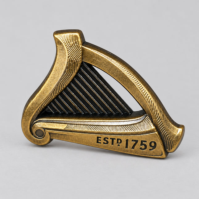 A small metallic harp-shaped Guinness pin or ornament in an antique gold/brass finish, resting on a speckled gray countertop with a blurred warm-toned background. The front is engraved with the text 'ESTD 1759'.