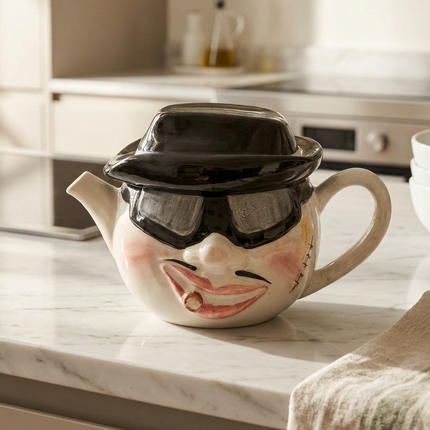 A ceramic teapot with a hand-painted face wearing a black bowler hat and dark sunglasses, sitting on a white marble kitchen countertop.