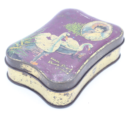 A vintage rectangular metal tin with rounded corners, featuring a purple lid decorated with a swan illustration and a circular portrait, with a distressed cream-colored base.