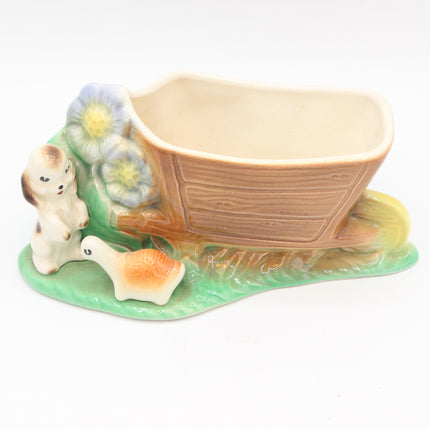 A vintage ceramic planter shaped like a small boat on a green grassy base, decorated with a rabbit, duck, and blue flowers.