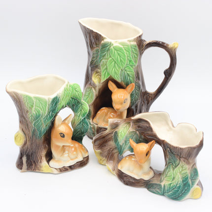 A ceramic three-piece woodland tableware set featuring a pitcher, a small bowl, and a small planter, all designed to look like tree stumps with green leaves and a small painted deer sitting in each piece.