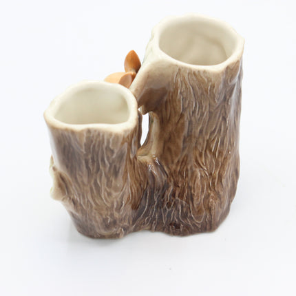 A ceramic vase designed to look like tree bark, featuring a textured brown and beige bark pattern with a small orange leaf detail, set against a plain white background.
