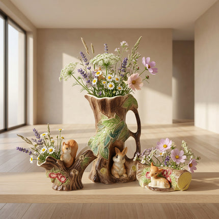 A ceramic pitcher and saucer set designed to look like tree trunks and branches, decorated with green leaves, small rabbits, and flowers, photographed on a white background.