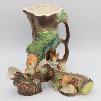 A ceramic woodland-themed pitcher set featuring a large tree-trunk-shaped pitcher with green leaf details, a small planter shaped like a log with a fox figurine, and a matching shallow dish with a rabbit figurine, all on a light gray background.