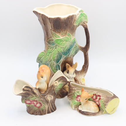 A ceramic pitcher and saucer set designed to look like tree trunks and branches, decorated with green leaves, small rabbits, and flowers, photographed on a white background.