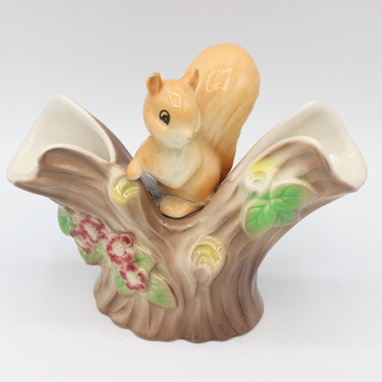 A ceramic figurine of a brown and beige squirrel perched on a stylized tree trunk base with painted flowers and leaves, photographed against a plain light gray background.