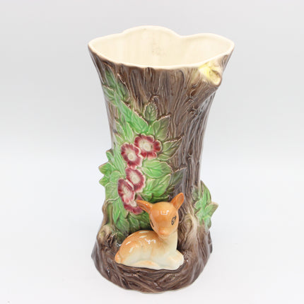 A ceramic vase shaped like a tree trunk with a small deer figurine at the base, green leaves, and pink flowers, shown on a light gray background.