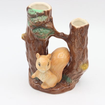 A ceramic squirrel figurine perched on a brown tree trunk base, with two small cup-like openings at the top of the trunk, shown on a plain white background.