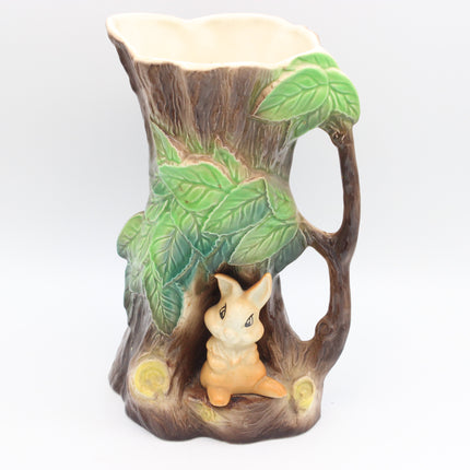A ceramic pitcher shaped like a tree stump with green leaves and a small rabbit figurine sitting in the base, shown on a plain light gray background.