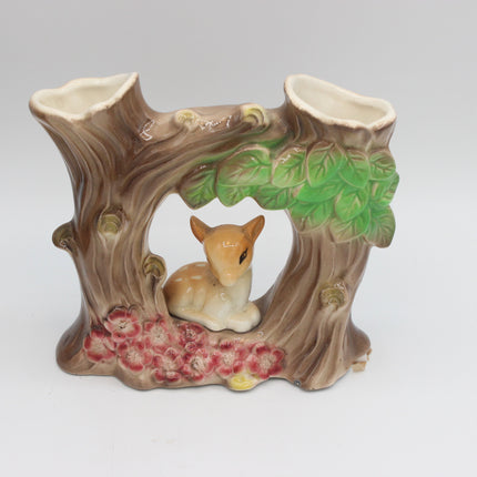 A ceramic woodland-themed candle holder shaped like a tree trunk arch with two candle slots at the top, a small deer figurine in the center, green leaves, and pink flowers at the base, photographed on a light gray studio background.