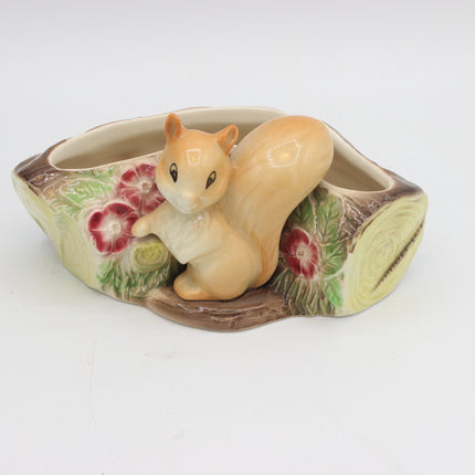 A ceramic planter shaped like a tree trunk with a glossy glazed finish, featuring a sculpted squirrel figure climbing on the front. The planter is decorated with painted red flowers and green leaves, and has a cream-colored interior.