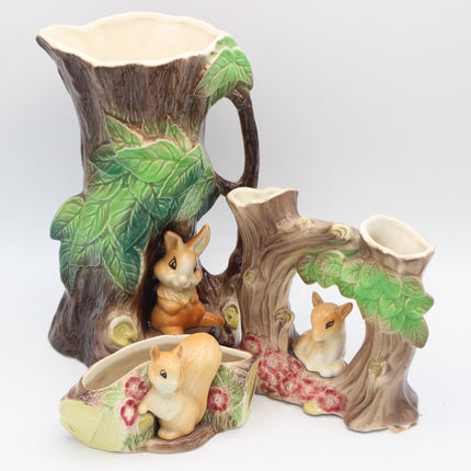 A ceramic woodland animal set shaped like tree stumps and logs, featuring green leaves, brown bark, and small rabbit and squirrel figurines, photographed on a light gray studio background.