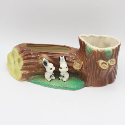 A ceramic woodland-themed planter shaped like a tree stump and log, with two small white rabbits sitting on a green patch in front. The planter has a brown bark-textured body with green leaf details and a cream-colored interior.