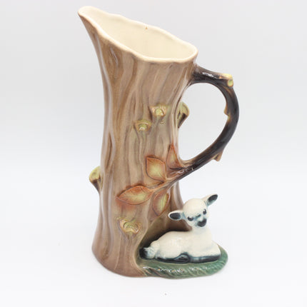 A ceramic pitcher shaped like a tree trunk with a curved handle, featuring painted leaves and a small white lamb figurine at the base, shown on a plain light gray background.