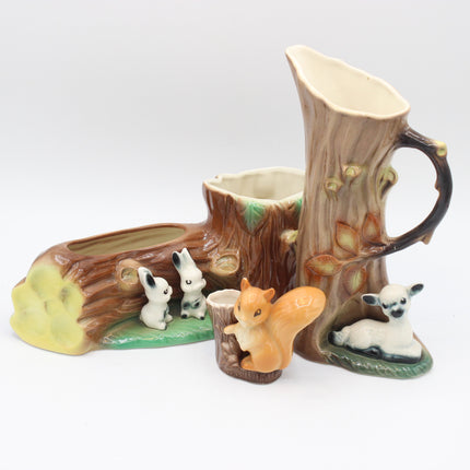 A ceramic woodland-themed pitcher and cup set shaped like tree stumps and trunks, with small animal figures including a rabbit, squirrel, lamb, and acorns, photographed on a light gray studio background.