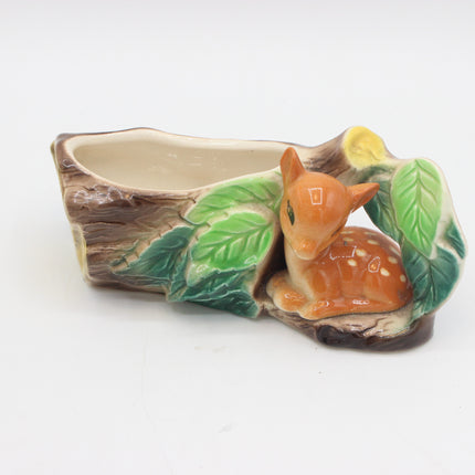 A ceramic planter shaped like a tree trunk with a small orange-brown fawn figurine nestled among green leaves on the right side, photographed on a plain white background.