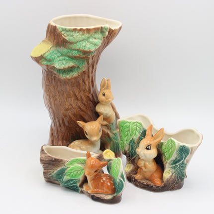 A ceramic woodland animal figurine set featuring a large tree stump with green leaves, three rabbits, and two deer in earthy brown, beige, and green glazes on a light gray background.