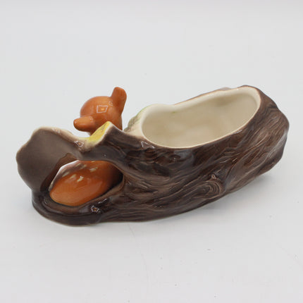 A ceramic planter shaped like a tree trunk with a small brown bear figurine and a peach-colored acorn, set against a plain white background.