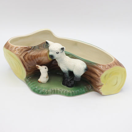 A ceramic planter shaped like a log with two small animal figurines, a white lamb and a white rabbit, on a green base, photographed on a white background.
