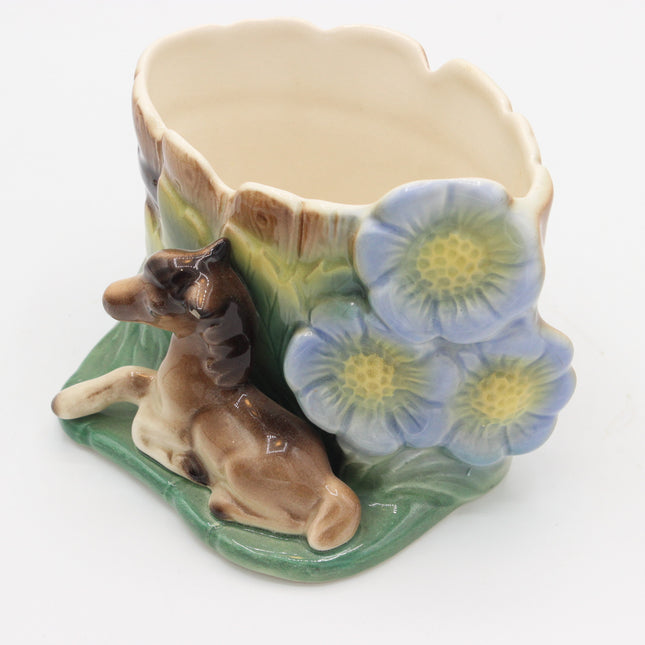 A ceramic planter shaped like a tree trunk with a brown dog figurine at the base and three blue flowers with yellow centers on the side, shown on a white background.