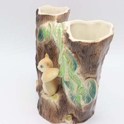 A ceramic double-vase holder designed to look like a tree trunk, featuring a small beige squirrel figurine and green leafy vine details, set against a plain light gray background.