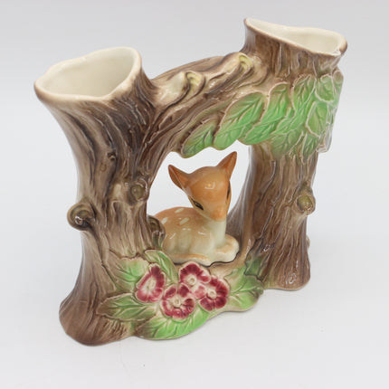 A ceramic woodland-themed candle holder shaped like a tree stump with a small deer figurine in the center and painted flowers and green leaves, shown on a plain light background.
