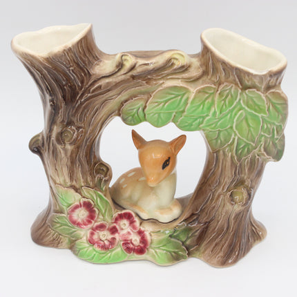 A ceramic woodland-themed candle holder shaped like a tree stump with a small deer figurine in the center, green leaves, and pink flowers, photographed against a plain white background.