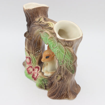 A ceramic bird feeder shaped like a tree stump with one open cup for a bird and a small cream-colored bird figurine nestled inside, decorated with green leaves and pink flowers on a white background.