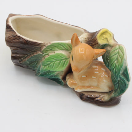 A ceramic planter shaped like a tree stump with a small glazed deer figurine nestled among painted green leaves, photographed on a plain white background.