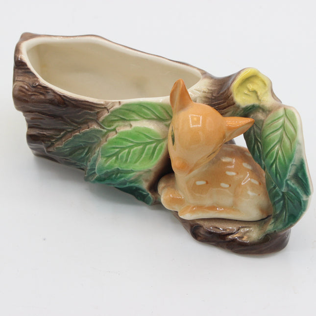 A ceramic planter shaped like a tree stump with a small glazed deer figurine nestled among painted green leaves, photographed on a plain white background.