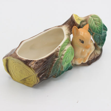 A ceramic planter shaped like a tree stump with a small orange-brown squirrel figurine nestled among green leaves on the side, photographed on a plain white background.