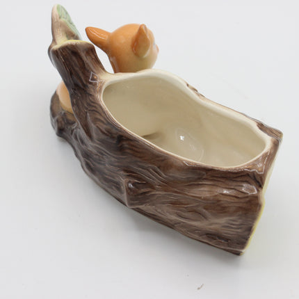 A ceramic decorative planter shaped like a tree trunk with a small orange animal figure peeking out from behind the top edge, photographed on a plain white background.